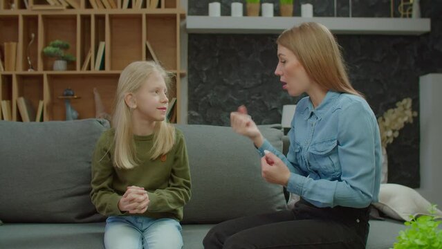 Cute preadolescent daughter with hearing loss and charming hearing impaired mother sitting on couch, friendly chatting with sign language and bonding while happy deaf family enjoying leisure at home.