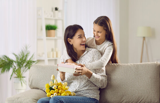 Caring small teen girl child make surprise to excited young mom congratulate with mothers day. Loving little daughter greeting mum give present and flowers to happy parent. Gratitude concept.