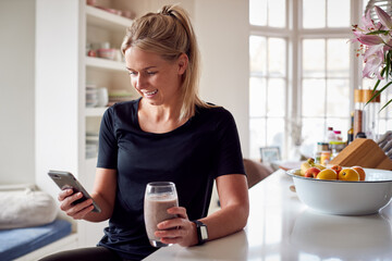 Woman With Healthy Shake In Kitchen At Home After Exercise Looking At Fitness App On Phone