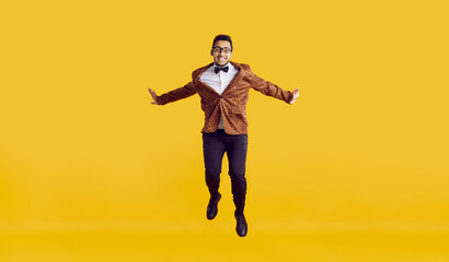 Fototapeta na wymiar Happy funny black man jumping in the studio. Cheerful young ethnic guy in a leopard jacket, bowtie and glasses jumping high in the air and smiling isolated on a vibrant yellow colour background