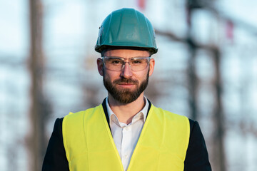 Fototapeta premium Electric engineer wearing helmet suit and safety vest checking datas on tablet with substation in background