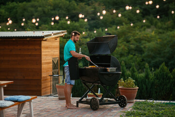 Young man grilling meat at barbecue outdoors with the help of barbecue gripper. - obrazy, fototapety, plakaty