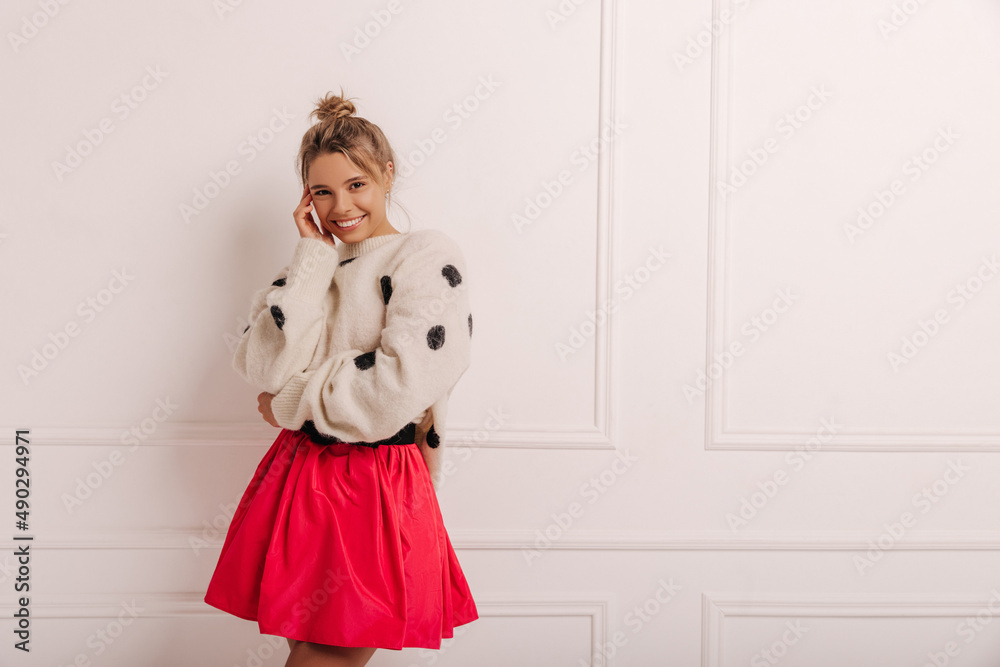 Wall mural Modest young european girl with bundle on her head smiles cutely at camera near white wall. Blonde in sweater and skirt. Concept of comfort, tenderness and warmth.