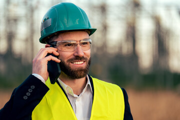 Electric engineer wearing helmet suit and safety vest making phone call with substation in background