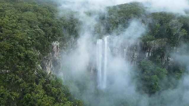 Mist covered mountain reveals a majestic tropical waterfall cascading down a cliff face. Cinematic drone view