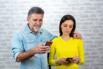 Photo of positive excited people man and woman screaming and looking at each other while both using mobile phones isolated over grey brick wall background.