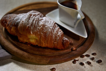 Breakfast concept with croissant, coffee in a white faience cup on a wooden tray and scattered coffee beans on the table. Color, full frame.