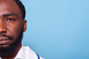 Cropped image of african american medic looking confident and relaxed in white lab coat with stethoscope on blue background. Half face closeup portrait of medical doctor with brown eyes.