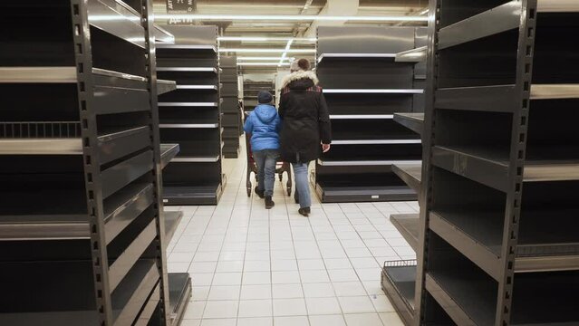 Mom and son in jackets and hats in mask walk with cart past empty shelves of store. Child looks at woman. View of customers from back. Crisis, default, complete sale of assortment of the store.