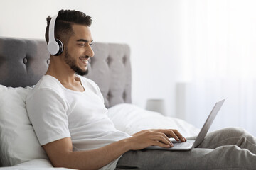 Young Arab Man Wearing Wireless Headphones Using Laptop While Relaxing In Bed