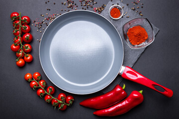 Empty ceramic pan with fresh vegetables in red color around: tomatoes and peppers. Grey kitchenware with a red handle on the dark background.