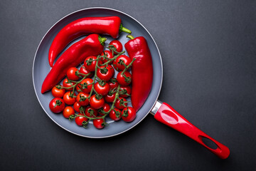 Ceramic pan full of fresh vegetables in red color: tomatoes and peppers. Kitchenware with a red handle on the dark background.