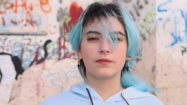 Serious Teenager in light blue oversize hoodie. Blue haired teen girl staying outdoor against graffiti wall and looking into camera. Head shot