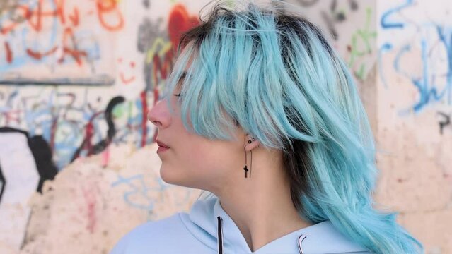 Serious Teenager in light blue oversize hoodie turn left. Blue haired teen girl staying outdoor against graffiti wall and looking into camera. Head shot