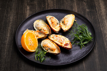 baked mussels with cheese, on a dark plate with herbs and lemon on a wooden background