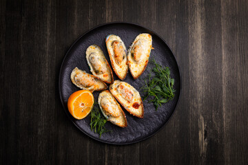 baked mussels with cheese, on a dark plate with herbs and lemon on a wooden background