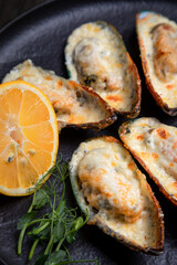 baked mussels with cheese, on a dark plate with herbs and lemon on a wooden background