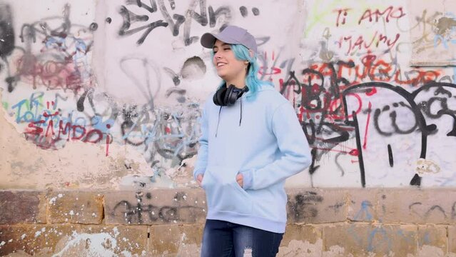 Teenager in light blue oversize hoodie wearing headphones and dancing. Blue haired teen girl stays outdoor against graffiti wall. Clothing mockup