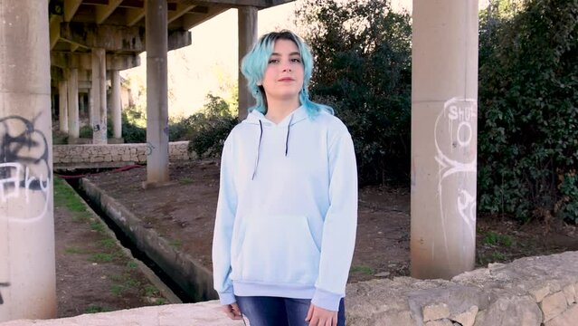 Smiling teenager wearing hoodie and staying against bridge pillars. Blue haired teen girl stays outdoors against bridge pillars. Clothing mockup