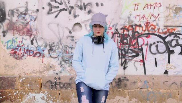 Teenager in light blue oversize hoodie with wireless headphones. Blue haired teen girl stays outdoor against graffiti wall. Clothing mockup