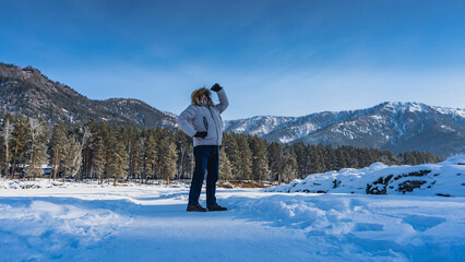 The frozen river is covered with snow. On the path between the snowdrifts there is a man in a down...