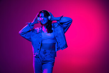 Music. Beautiful young emotional girl, student with long dark hair isolated on gradient pink-blue background in neon light. Concept of emotions, youth