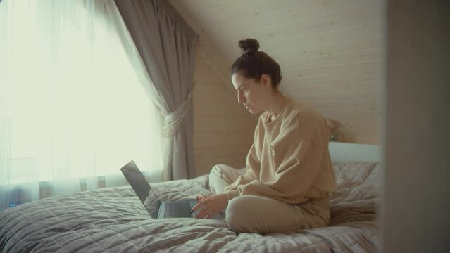 Concentrated woman works on laptop. Female freelancer uses modern PC. Lady rests on bed with notebook. Female blogger home workplace. 4K footage