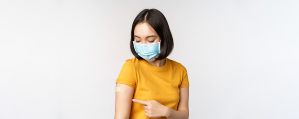 Covid-19, vaccination and healthcare concept. Cute asian girl in medical face mask, showing band...