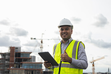 Asian engineer handsome man or architect use tablet with white safety helmet in city construction site . Standing on rooftop building construction at capital.