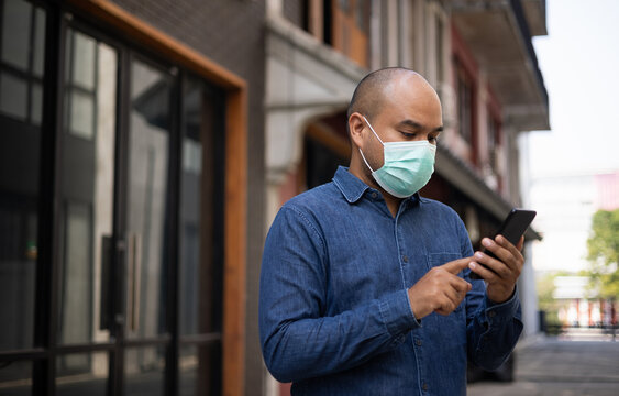 Young indian using smartphone standing in front of office building. Asian man wearing protective mask during pandemic.