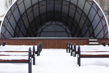 Naklejka na ściany i meble A dark wooden and metal stage in the park with benches covered with snow. A small amphitheater. Snowy winter