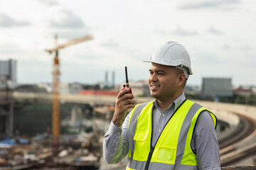 Asian engineer handsome man or architect use radio communication with white safety helmet in city construction site . Standing on rooftop building construction at capital.