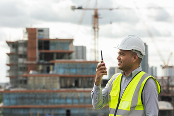 Asian engineer handsome man or architect use radio communication with white safety helmet in city construction site . Standing on rooftop building construction at capital.