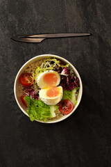 Healthy bowl with poached egg, bulgur, tomatoes and lettuce. on a dark background in an eco paper box. Healthy food delivery concept.