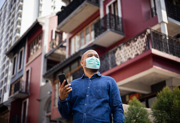 Obraz na płótnie Canvas Young indian using smartphone standing in front of office building. Asian man wearing protective mask during pandemic.