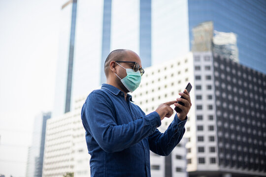 Young indian using smartphone standing in front of office building. Asian man wearing protective mask during pandemic.
