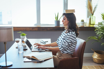 Portrait of a young attractive woman at the desk with books on her head, sitting straight, working...