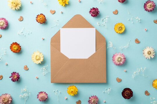 Top view photo of big open craft envelope paper card inside and many different cute flowers with white gypsophila branches wooden confetti in shape of hearts scattered on the pastel blue background