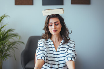 Shot of young attractive woman at the desk with books on her head while working with computer at...
