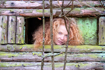 portrait of a beautiful sexy lovely mature redhead woman with long red curly hair, looking out of window of wooden shed, hunter high seat, in forest