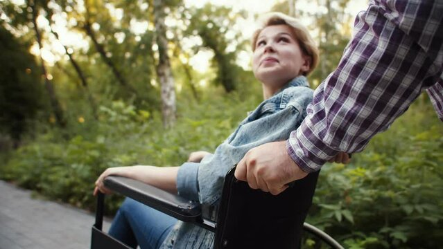 Disabled woman in wheelchair that her boyfriend is pushing, talking to him while they are walking in park, side view. Man is pushing wheelchair on park with disabled woman