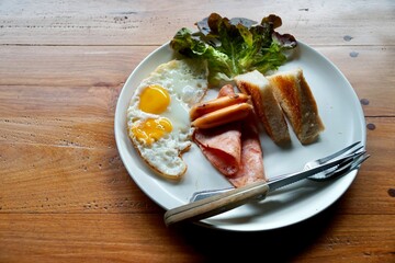 top view of breakfast set. fried eggs, sausages, ham sliced, bread and red oak on white dish with fork and knife for serve. wooden table as background in morning light with copy space.