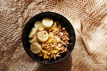 Cornflakes topping with banana and almonds nut for eating with milk or yoghurt in black bowl placed on brown placemat and wooden spoon. Ready to eat for breakfast or before workout. Healthy food.