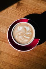 Cappuccino with latte art in a red ceramic cup on a saucer on a wooden table.