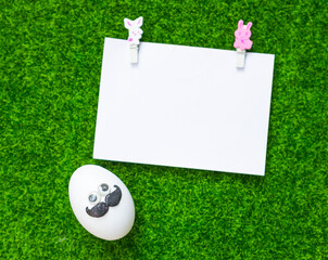 Naklejka premium Funny man face Easter egg with blank paper card on green grass background, simplae easter, blank card with funny egg