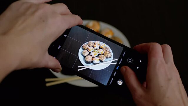 Female Hands Taking Photos of Prepared Food on a Smartphone on Table. Girl blogger shoots a picture, a photo, video review of sushi rolls on a mobile phone camera. Photographing food. Home kitchen.