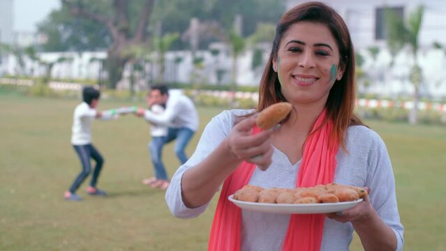 A beautiful housewife wearing a white Kurti and Chunni offers traditional Gujiya - Holi celebrations  an Indian sweet  a tasty cuisine. A young man and his little kids enjoy playing with a water gu...