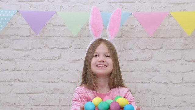 Cute little girl smiling giving basket with colorful eggs on Easter. Happy child wearing bunny rabbit ears with present. Spring holidays