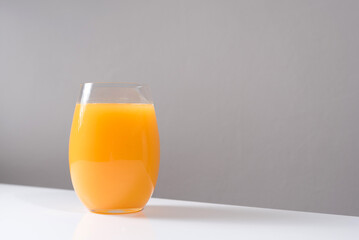 Un vaso de zumo de naranja exprimido en un vaso de cristal con trozos de naranja sobre una mesa blanca con un fondo gris