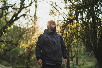 Chico con barba y bajito sobre una roca con el sol de fondo en un bosque haciendo senderismo